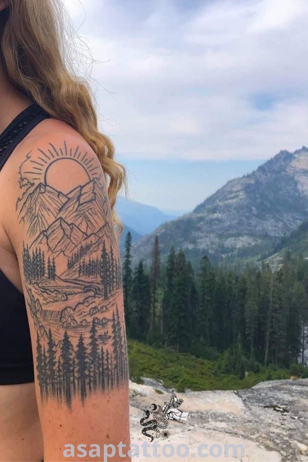 A woman with a tattoo on her arm looks at the mountains and trees in the distance, showcasing beautiful Western tattoos ideas like cowgirl and nature tattoo sleeves. Discover more inspiring tattoos at asaptattoo.com.