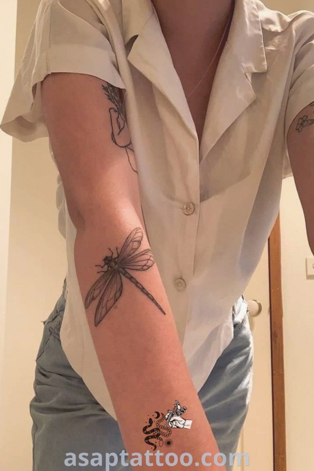 A woman takes a selfie in the mirror, showcasing a detailed dragonfly tattoo on her arm. This stunning design highlights artistic features that will inspire you to explore dragonfly tattoo ideas at asaptattoo.com. Ideal for those seeking meaningful ink, this tattoo is a gorgeous and elegant choice.