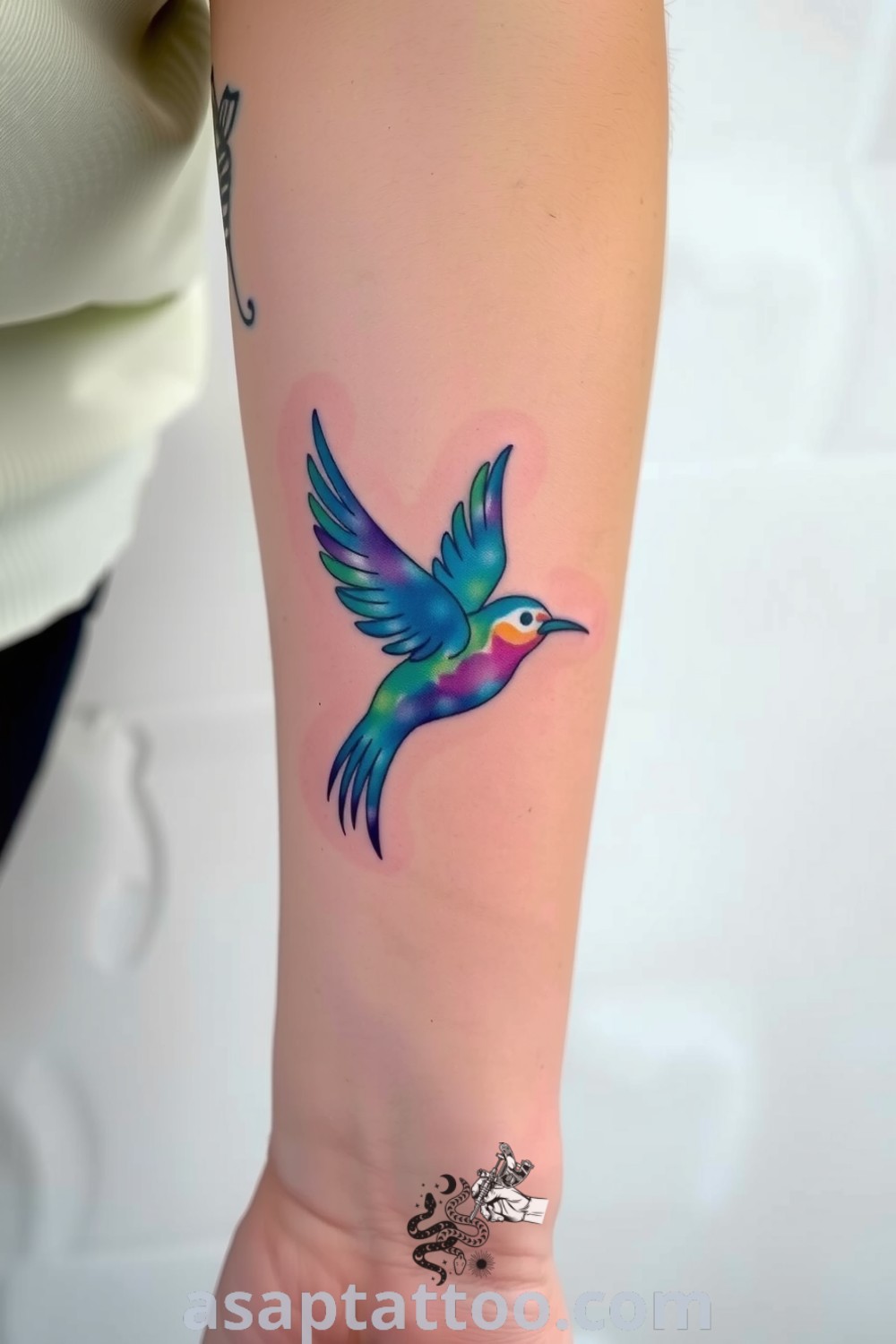 Watercolor bird tattoo in vibrant blues, purples, and greens depicting a bird in flight with delicate brushstrokes. A gorgeous tattoo idea that symbolizes freedom and creativity, perfect for body decoration. asaptattoo.com