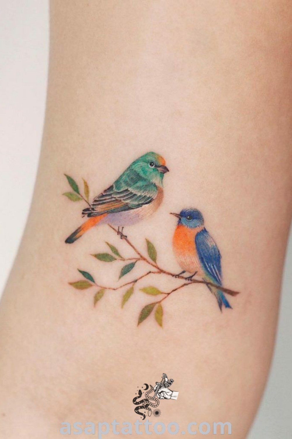 Tattoo of two small birds on a branch, one blue and the other orange, embellished with leaves around their ankles. A gorgeous design for lovers of colorful bird tattoos, offering inspiring ideas that you will absolutely love. Discover more at asaptattoo.com.
