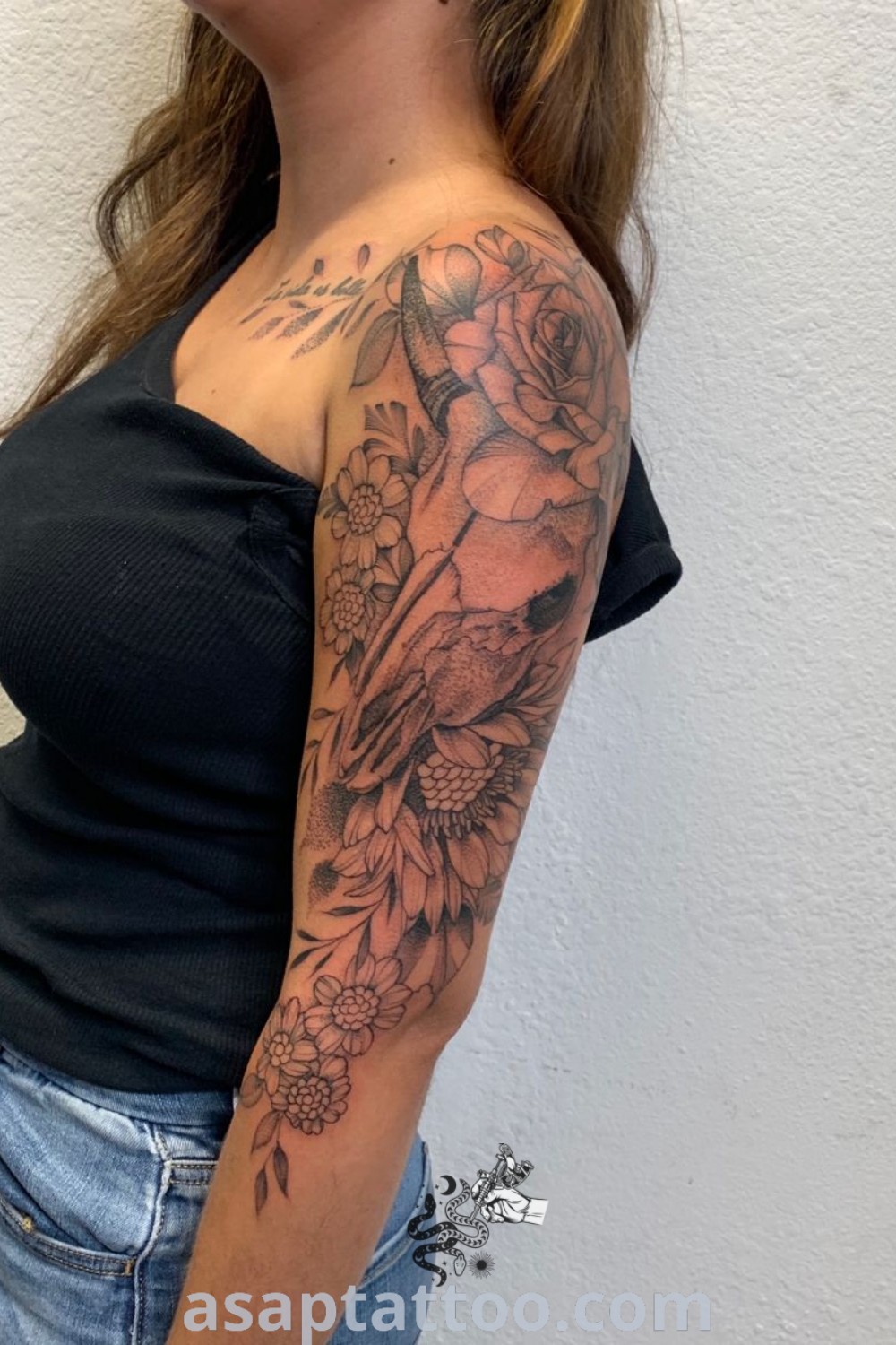 A woman with a beautiful Western tattoo on her arm and shoulder stands in front of a white wall, showcasing inspiring bull skull and cow skull designs. Discover amazing tattoo ideas for females at asaptattoo.com.